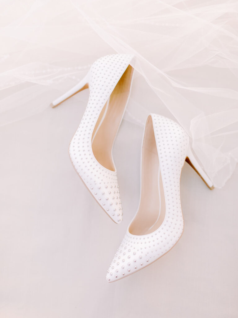 Elegant white high heels adorned with sparkling rhinestones, set against a soft backdrop of sheer white fabric.