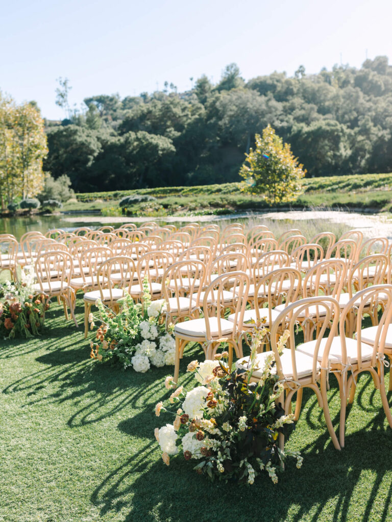 Rows of elegant wooden chairs are arranged outdoors on a grassy lawn, surrounded by lush greenery and flower arrangements, under a clear blue sky.