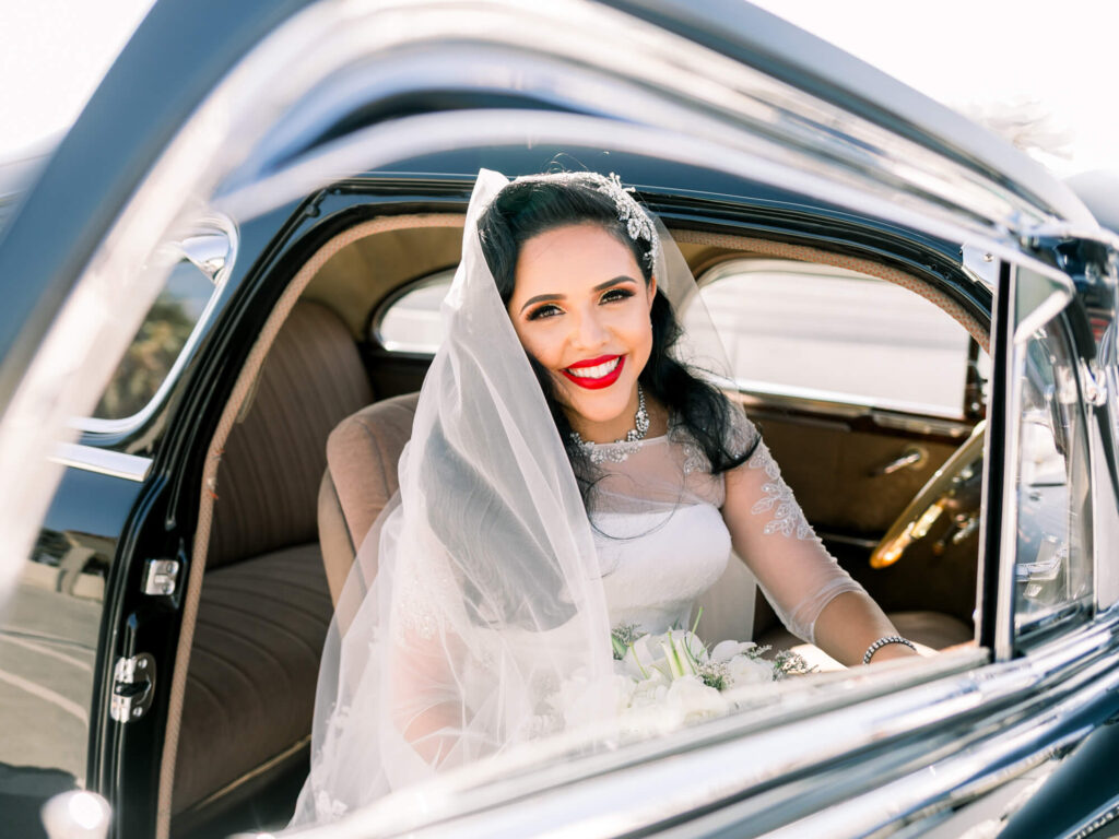 A joyful bride with red lipstick smiles from the window of a vintage car. She wears an elegant white dress and veil.