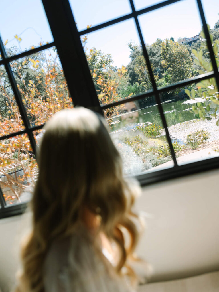 A woman with long blonde hair gazes out a window at a serene landscape with a pond and autumn trees.