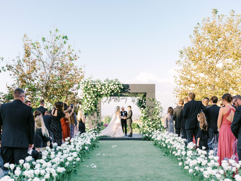 A picturesque outdoor wedding ceremony with a couple standing under a floral arch. Guests in formal attire are seated along an aisle lined with white roses.