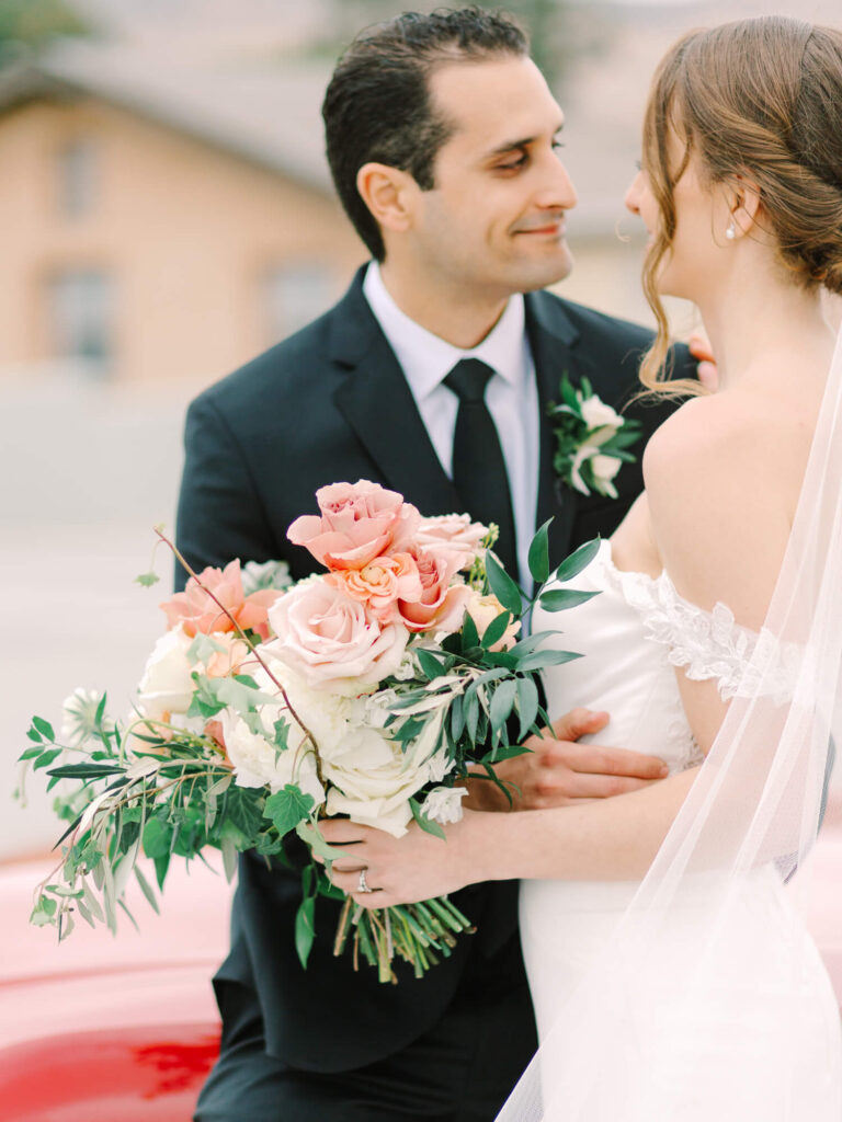 Bride and groom lovingly gaze at each other. Bride holds a bouquet of pink and white roses. Groom in dark suit and tie.