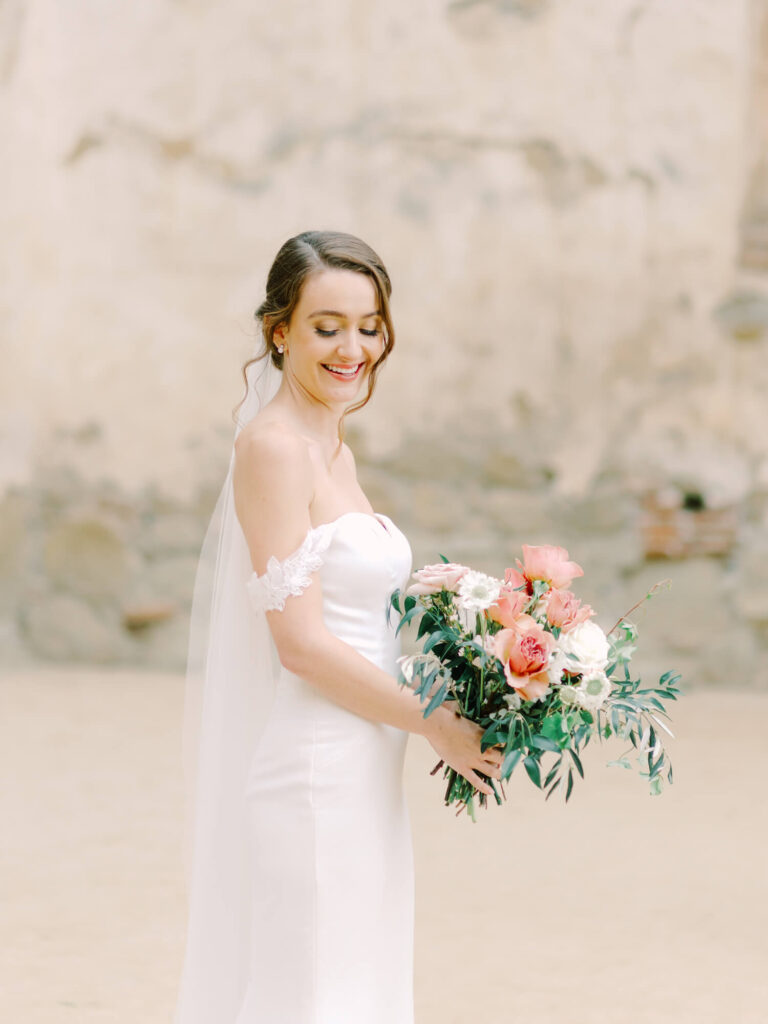 Bride in off-shoulder white gown, smiling while holding a colorful bouquet with pink and white flowers.