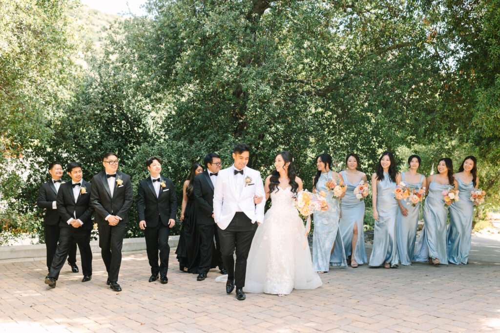 A joyful wedding party walking outdoors, fronted by the bride in a white gown and groom in a white jacket. Bridesmaids in blue and groomsmen in black follow.
