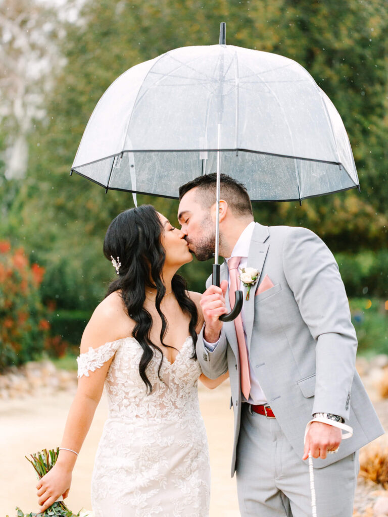 A bride in a lace dress and groom in a gray suit share a kiss under a clear umbrella on a rainy day.