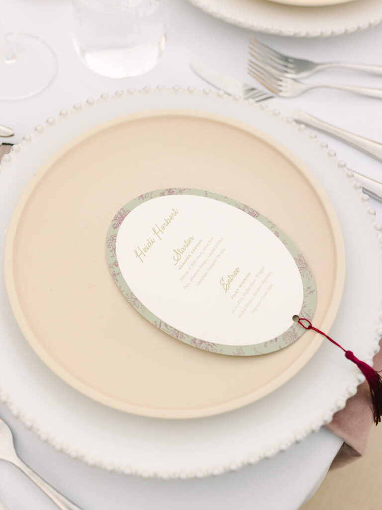 Elegant table setting with a menu card on a beige plate, surrounded by cutlery and a glass.
