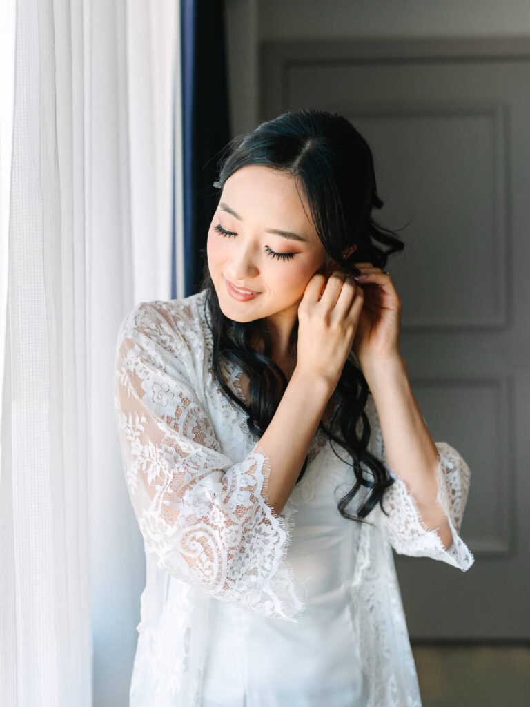 A woman stands by a sunlit window, smiling softly as she adjusts her earring. She wears a delicate, white lace robe.