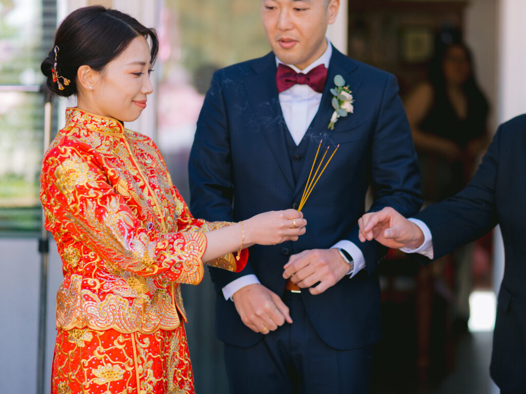 A couple in traditional attire participates in a ritual. The woman wears a red and gold ornate dress, holding incense. The man is in a suit with a bow tie, looking focused.