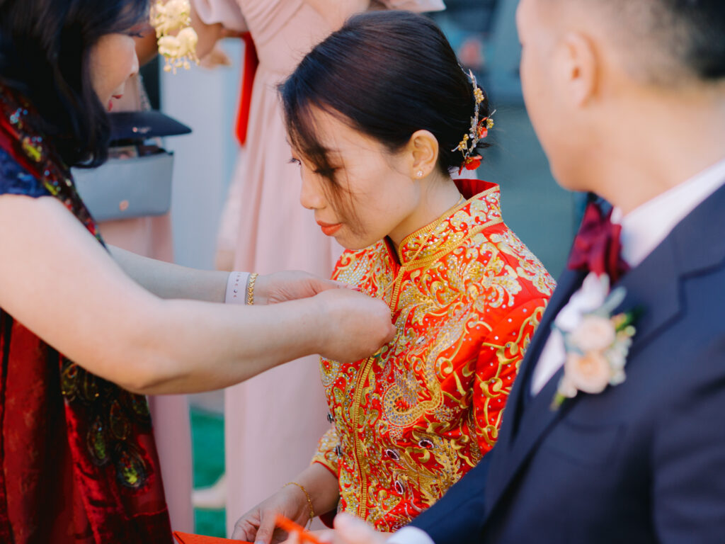 A woman in an intricate red and gold traditional gown is being gifted a gold necklace by another person.
