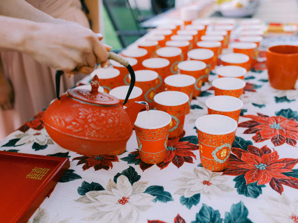 A red teapot with floral details pours tea into paper cups, arranged in rows on a poinsettia-patterned tablecloth.