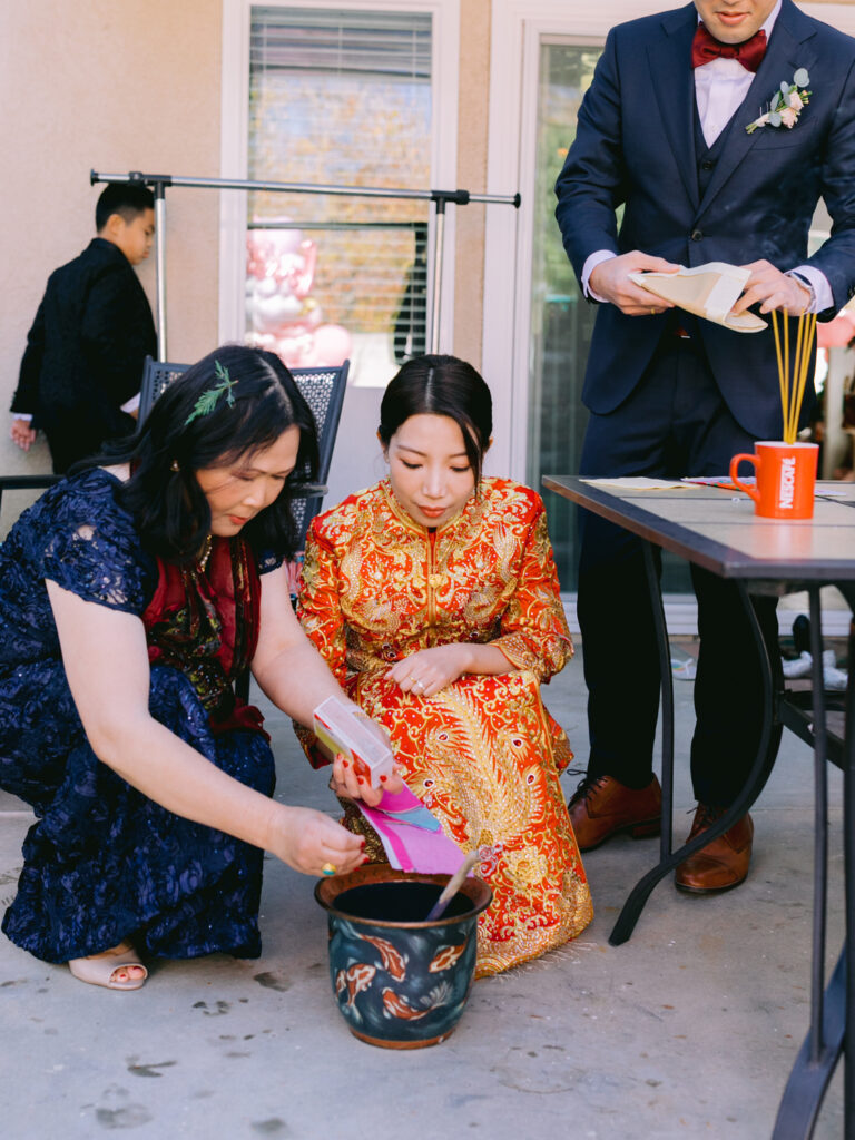 A woman in a blue dress and another in a red traditional dress are engaged in a ritual, burning paper in a pot.