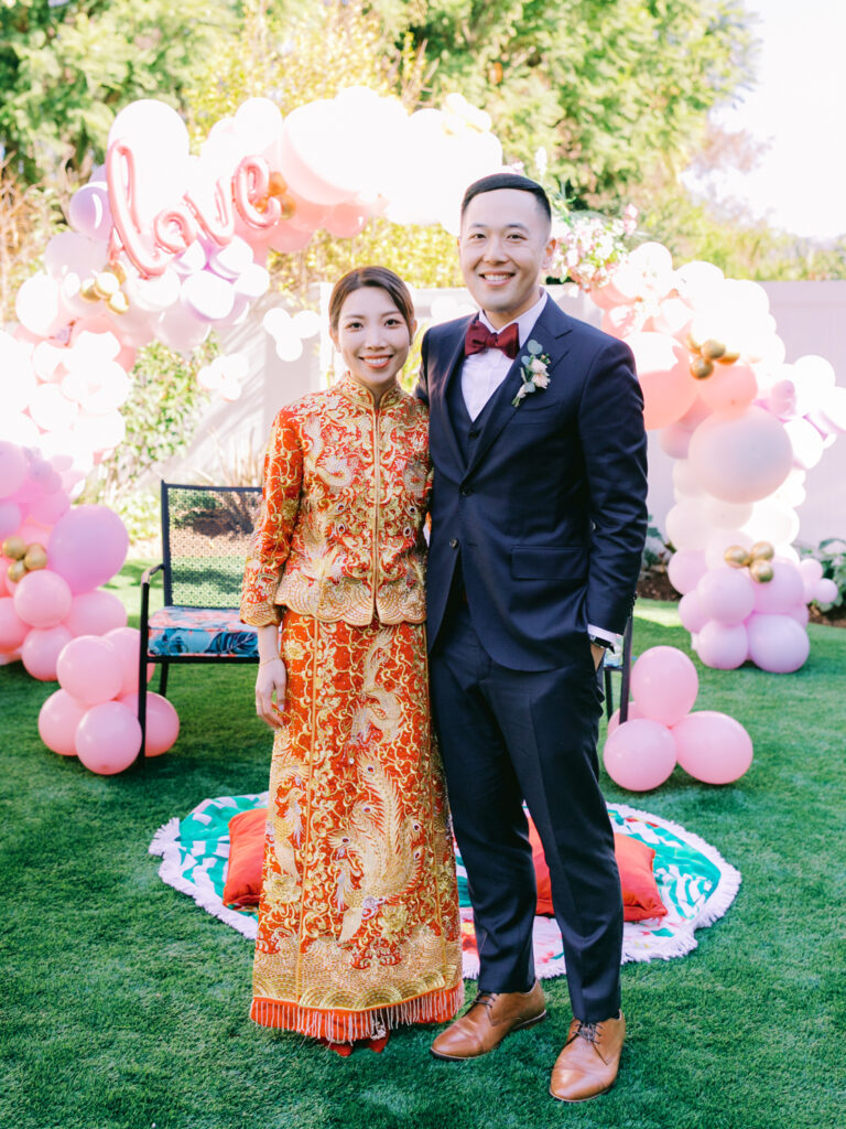 A couple stands smiling outdoors in formal attire, surrounded by pink and white balloons with the word "love."