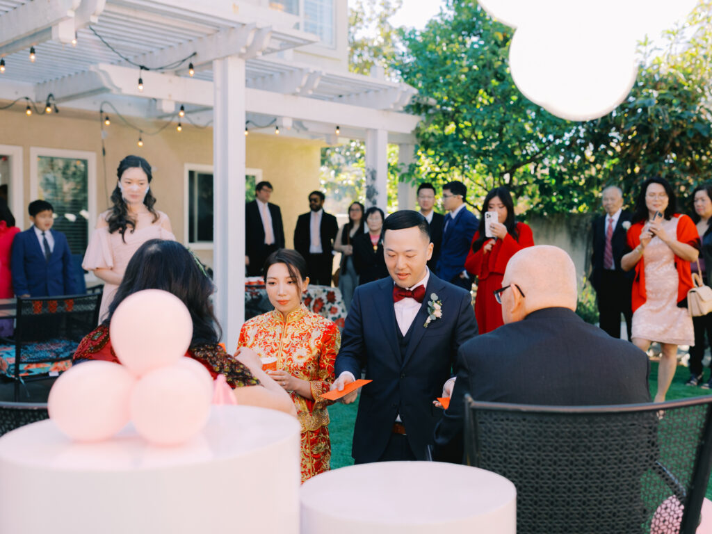 A couple in formal attire participates in a tea ceremony outdoors, surrounded by guests. The woman wears a red dress and the man a suit with a bow tie