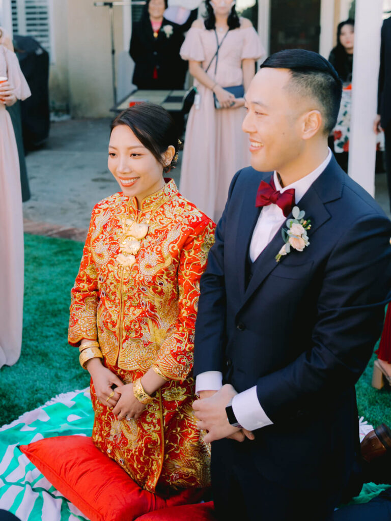 A couple kneels smiling during a traditional ceremony. The woman wears an ornate red and gold dress; the man wears a dark suit with a red bow tie.