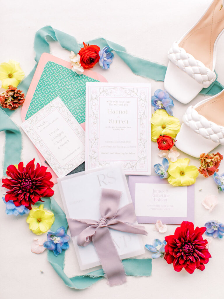 Wedding stationery featuring elegant invitations with floral designs, surrounded by colorful flowers, teal ribbons, and white braided sandals.