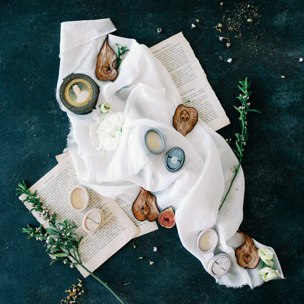 A dark textured background with vintage books, white fabric, dried fruits, and green sprigs. Vintage lockets rest on the fabric, adding an elegant touch.