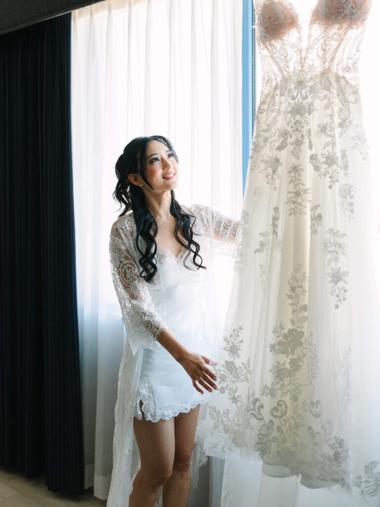 A woman in a lace robe admires a detailed wedding gown with floral patterns, holding it near a window.