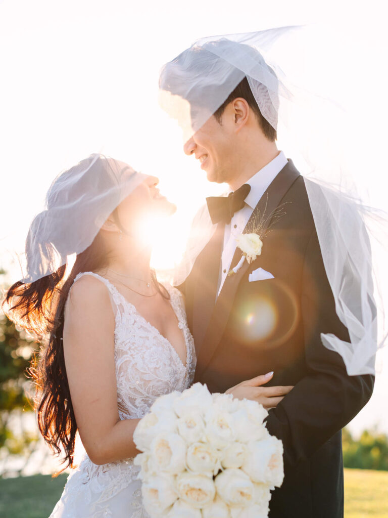 Bride and groom in wedding attire embrace joyfully, with veils blowing in sunlight. She holds white roses, and both smile warmly against a bright sky.