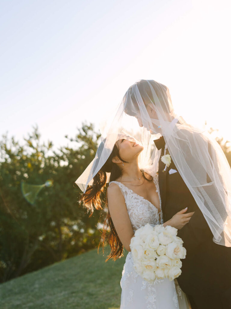 Bride and groom under a veil share an intimate moment outdoors, with the sun setting behind them.