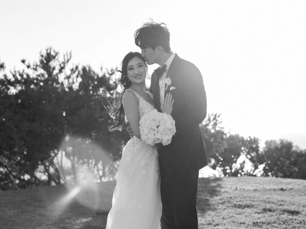 Black and white photo of a joyful couple on their wedding day. The groom kisses the bride's forehead while she holds a bouquet.