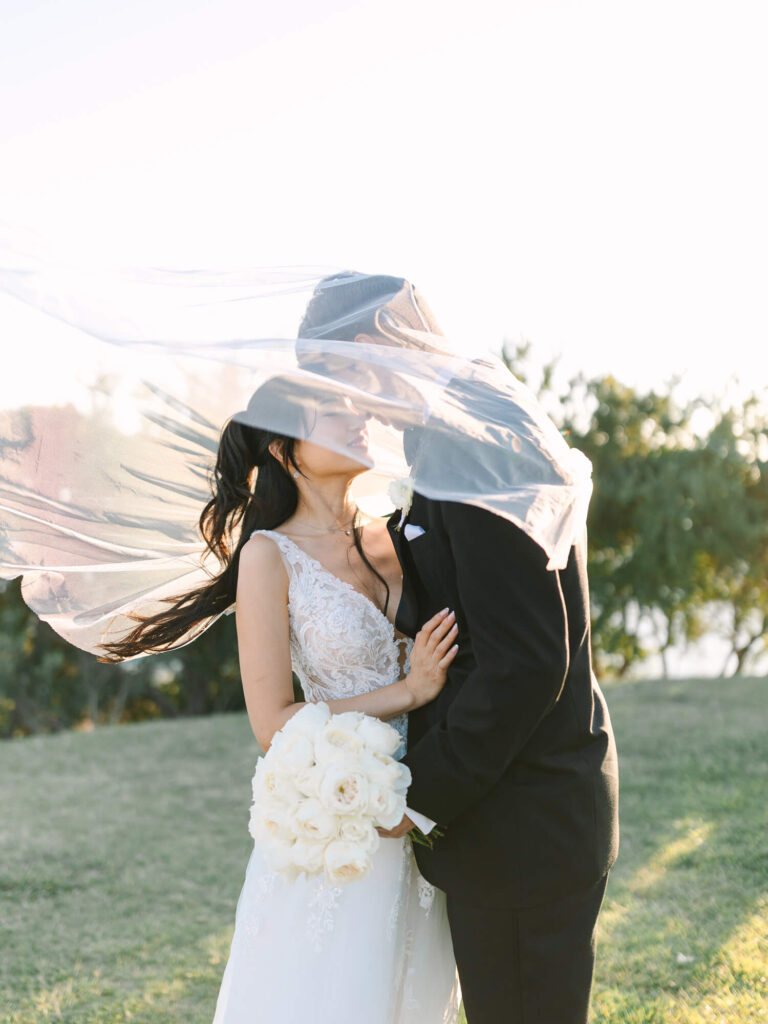 Bride and groom kiss outdoors, veils billowing in sunlight. Bride holds white rose bouquet, wearing lace dress.