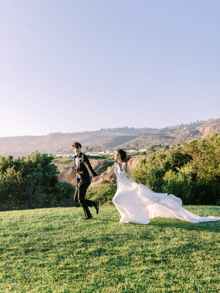 A bride in a flowing white dress and a groom in a black suit joyfully run hand in hand on lush grass, with a scenic hillside in the clear background.