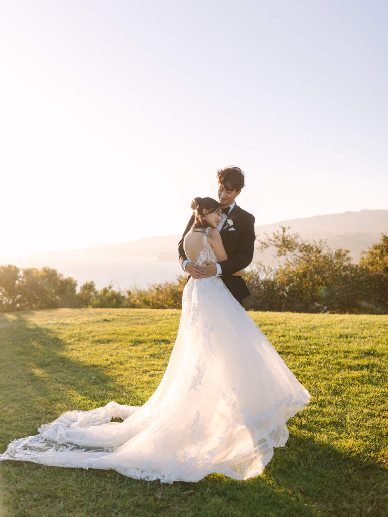 A bride and groom embrace lovingly on a sunlit grassy hill, her lace dress trailing behind.