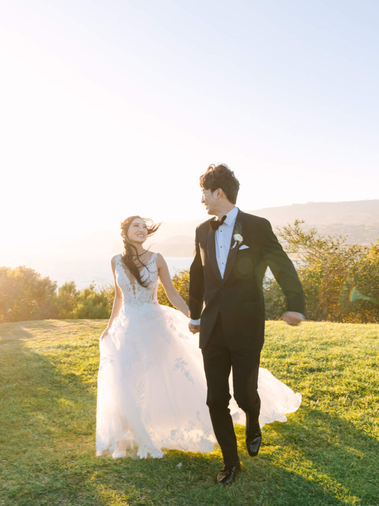 A joyful couple, dressed in wedding attire, holds hands while walking on sunlit grass. The bride's dress flows, and both smile warmly.