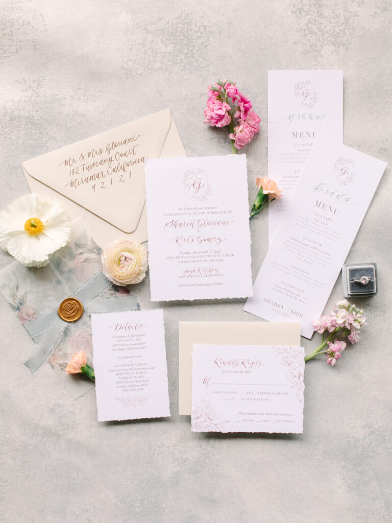 Elegant wedding invitation suite on textured background, featuring floral designs and calligraphy. Includes RSVP, menu cards, a wax-sealed envelope, flowers, and a ring box.