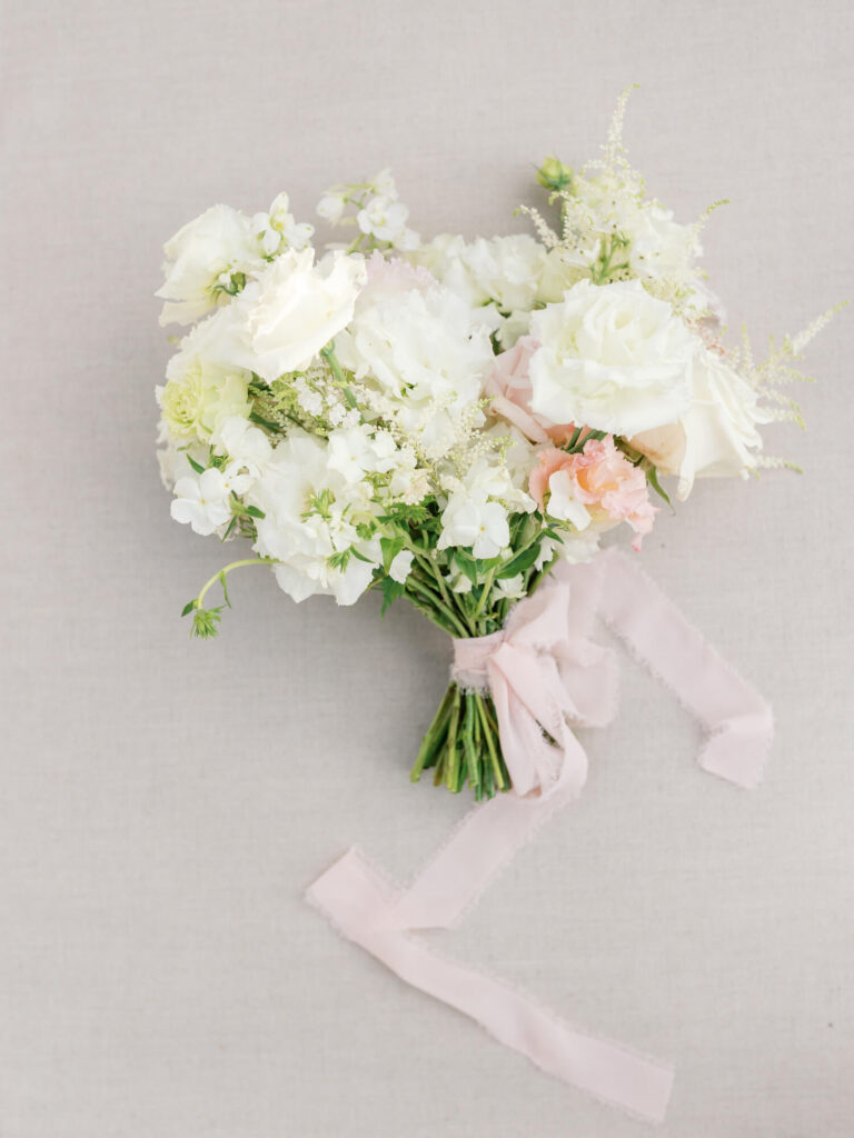 A delicate bouquet of white and pale pink flowers, including roses, tied with a soft pink ribbon. The arrangement lies on a light beige fabric background.