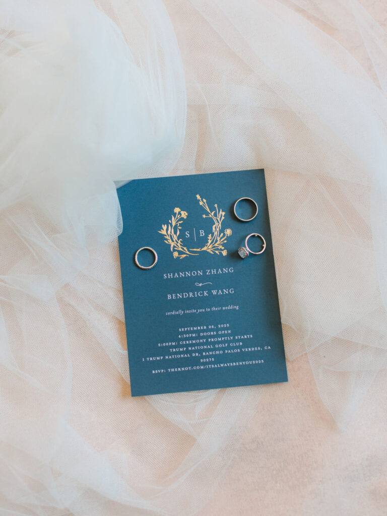 Blue wedding invitation with gold floral design and initials "SB." Surrounded by four silver rings, placed on a soft, white tulle fabric. 