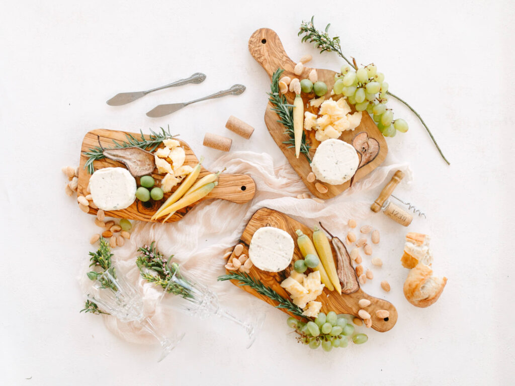 Three wooden boards with cheese, grapes, nuts, and herbs on a light fabric. Two knives and wine corks add an elegant touch.