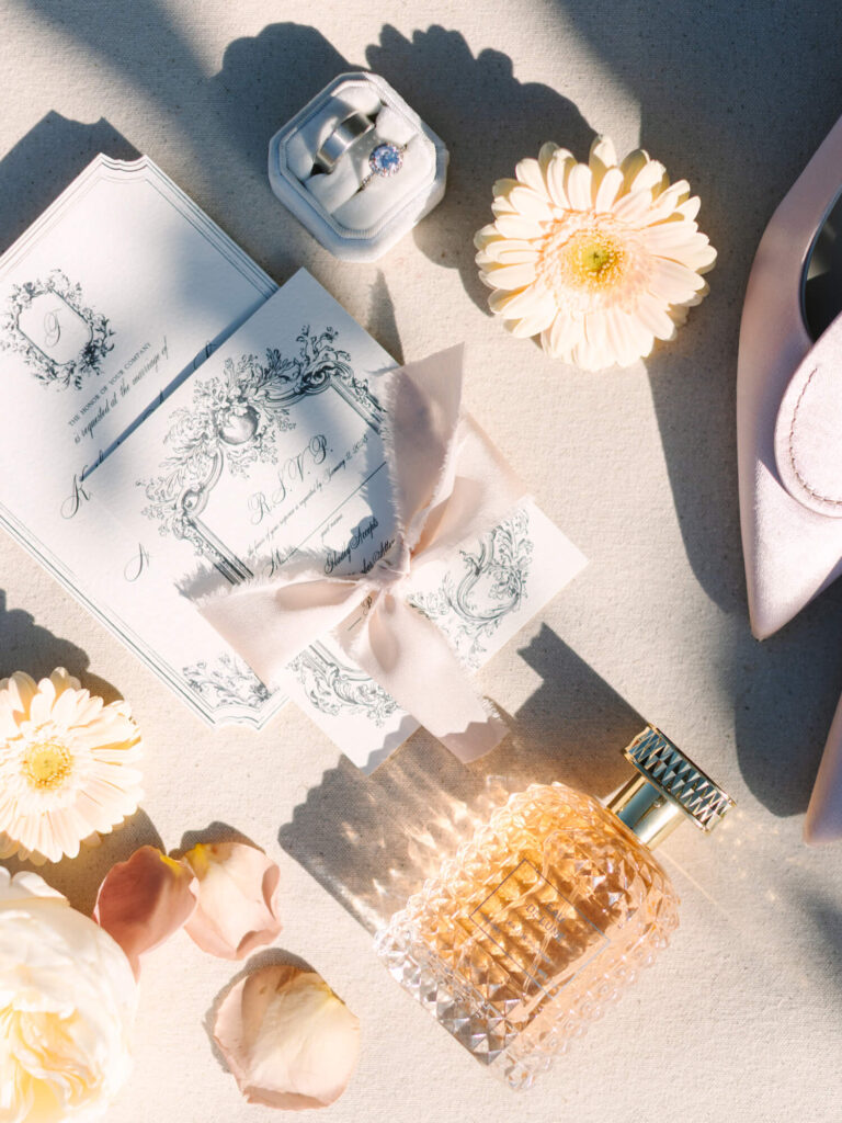 Elegant flat lay with an ornate invitation, ribbon, floral ring box, pink shoe tip, pale yellow flowers, rose petals, and a faceted perfume bottle.