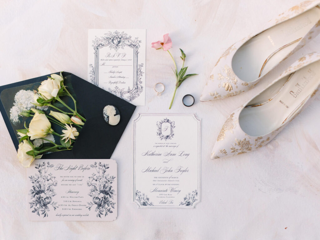 Elegant wedding flat lay with floral invitations, a black envelope with white flowers, two rings, a pink flower, and gold and white heels.