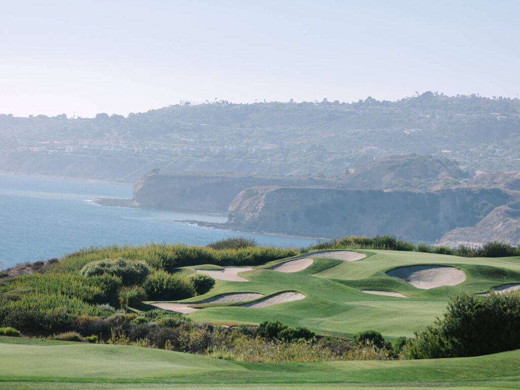 Coastal golf course with manicured greens and sand bunkers, set against a backdrop of ocean cliffs and hazy hills under a clear blue sky.