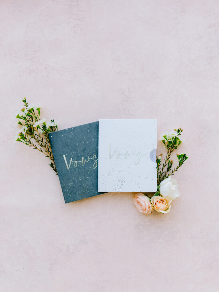 Two vow books, one dark and one light, are surrounded by sprigs of delicate flowers and roses on a soft pink background.