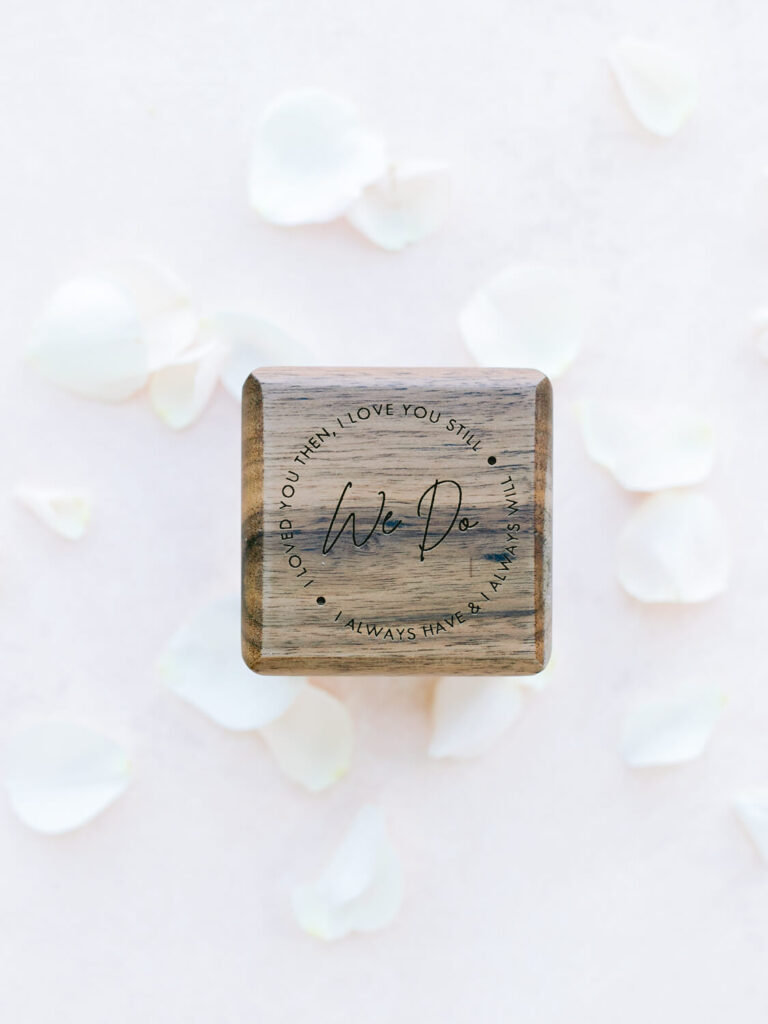Wooden box with "We Do" engraved, surrounded by text "I loved you then, I love you still, I always have, I always will." Set against scattered white petals.