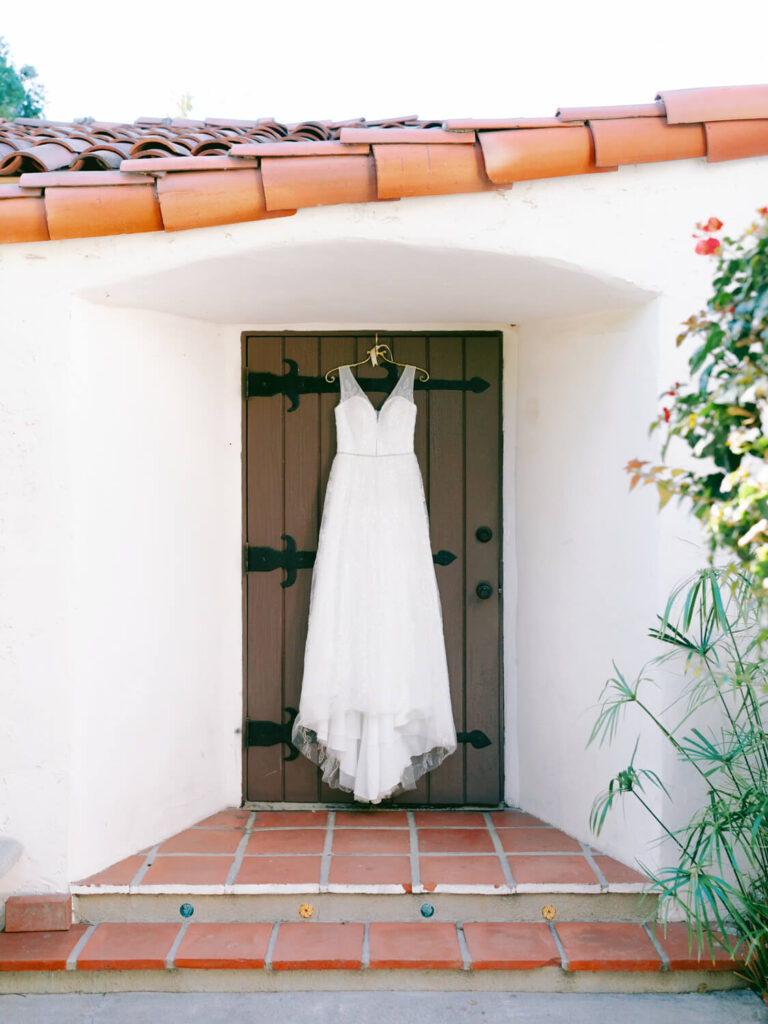 White wedding dress hangs on a rustic wooden door with terracotta tiles, set against a white stucco wall, surrounded by greenery.