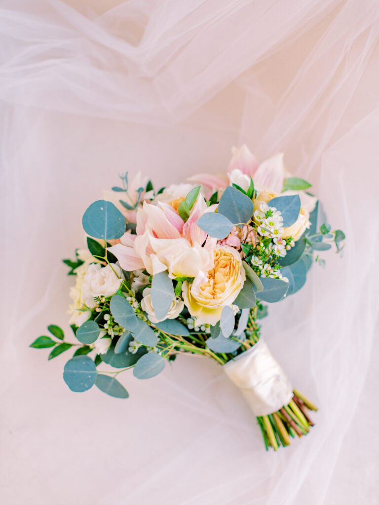 A delicate bridal bouquet with pink lilies, cream roses, white flowers, and eucalyptus leaves, wrapped in white ribbon, on soft white fabric.