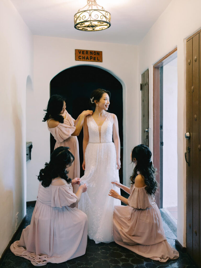Bride in a white gown stands in a chapel hallway, smiling as three bridesmaids in pink dresses adjust her dress.