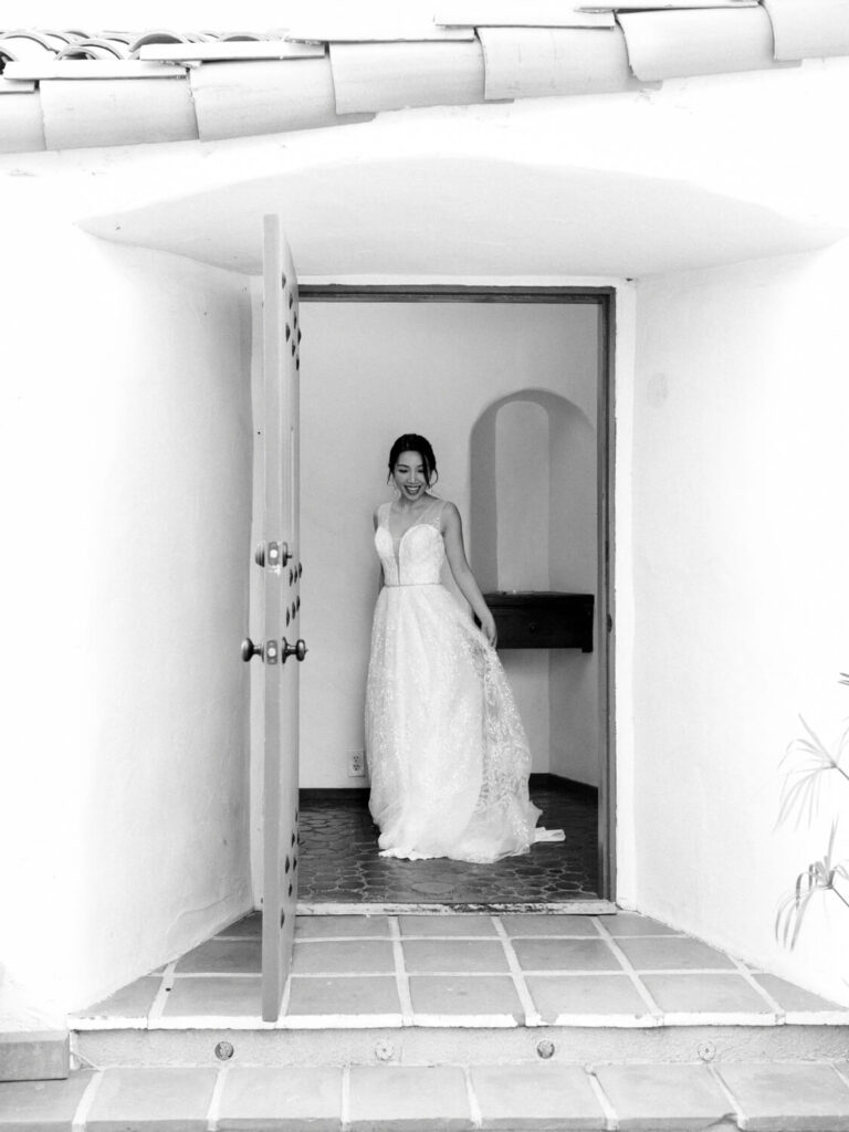 Bride in a strapless gown stands in an open doorway of a white archway, smiling warmly.