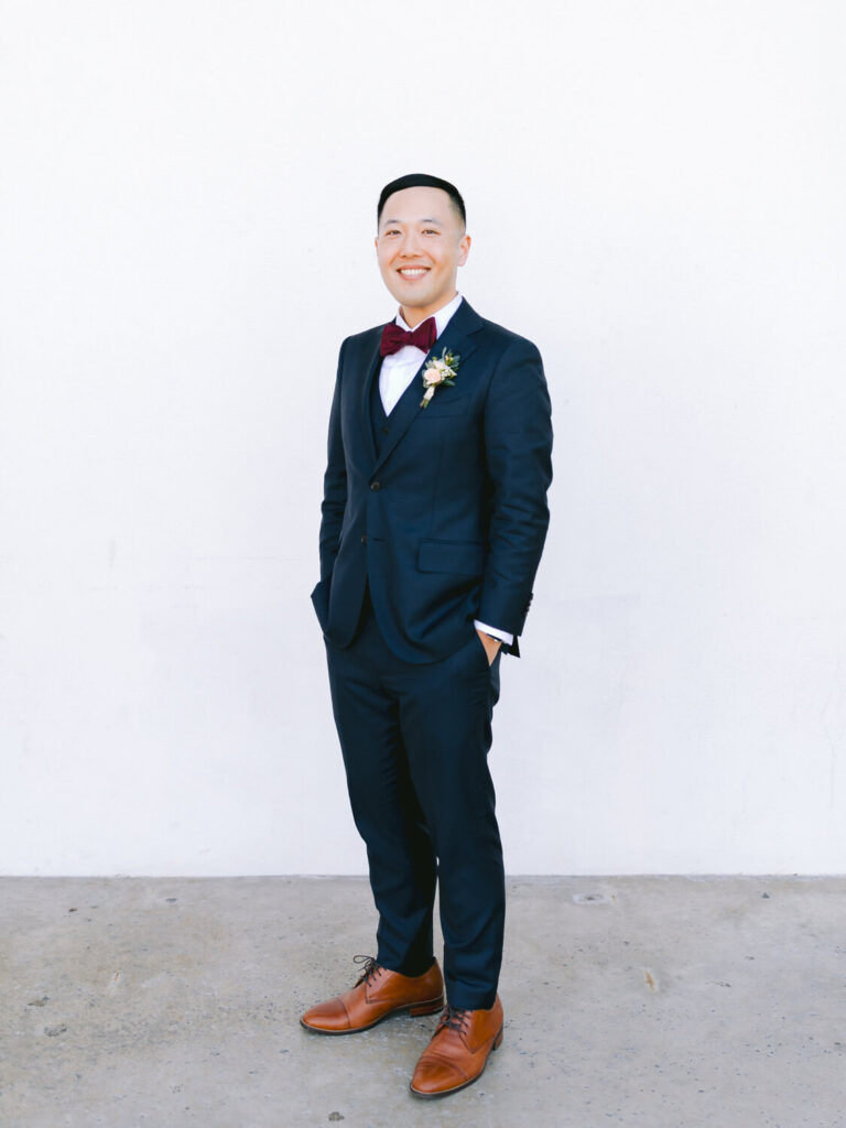 A person in a navy suit with a burgundy bow tie and brown shoes stands smiling against a plain white background.