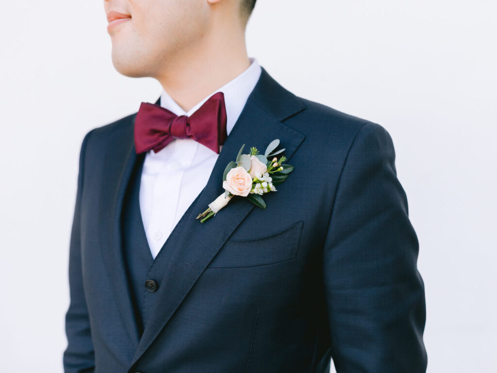 Man in a dark suit with a pale pink boutonniere and a burgundy bow tie stands in profile against a plain white background.