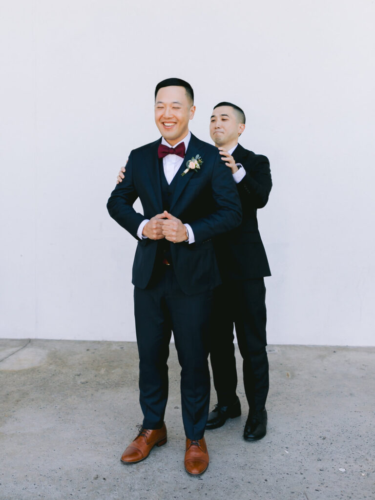 Two men in navy suits stand against a white background. The man in front, smiling and wearing a bow tie and boutonniere, is helped by his groomsman.