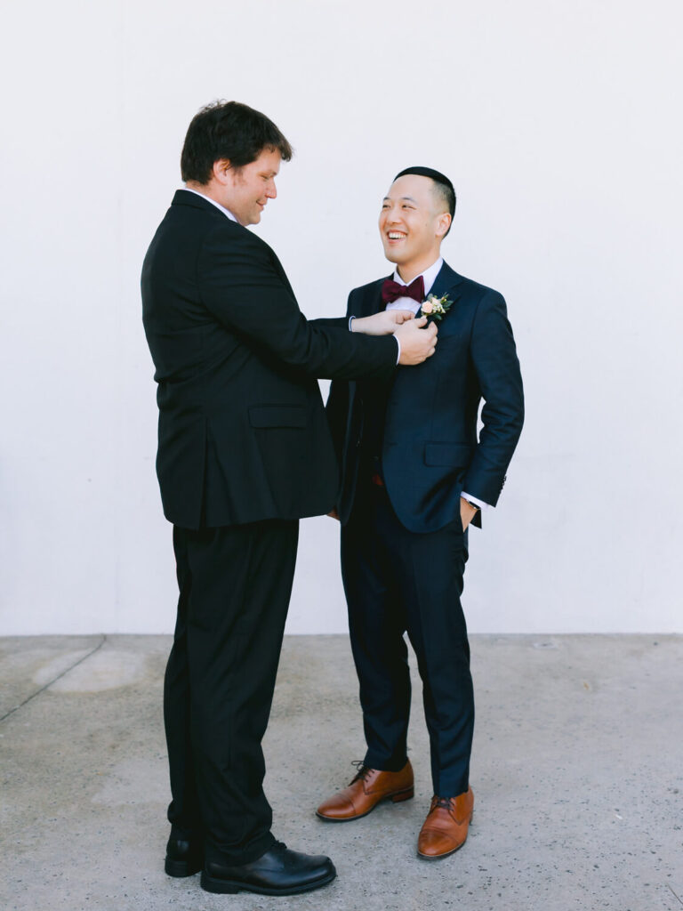 Two men in suits stand against a plain white background, smiling warmly. One adjusts the other's boutonniere.