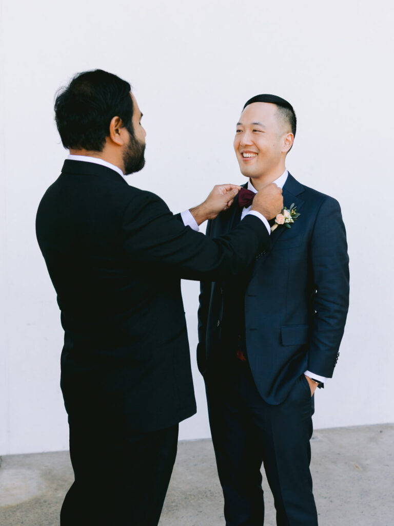 Two men in suits stand against a plain white background, smiling warmly. One adjusts the other's bowtie.