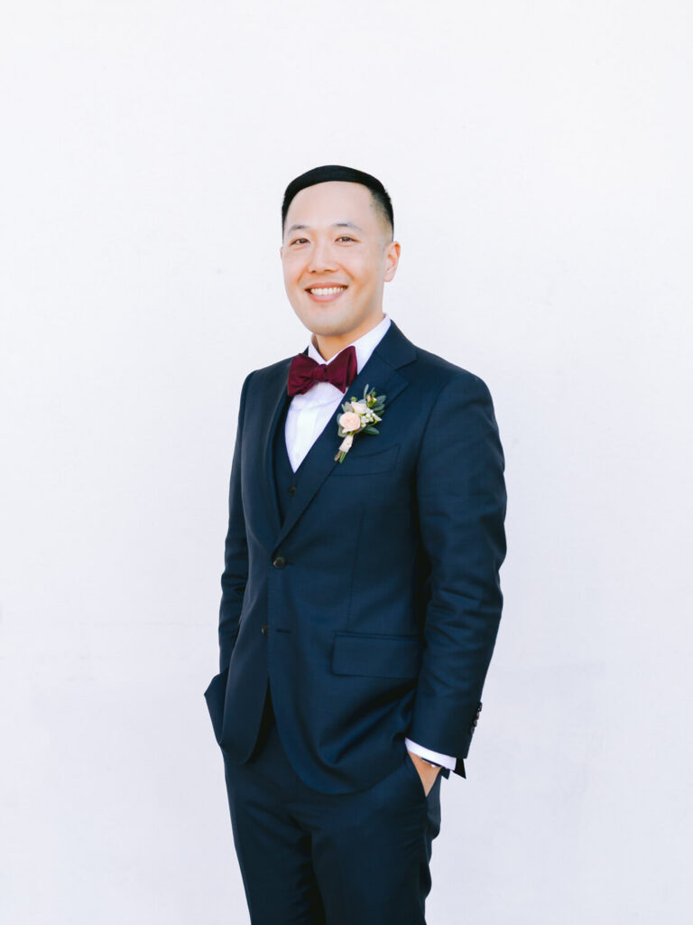 A groom in a navy suit with a burgundy bow tie and brown shoes stands smiling against a plain white background.