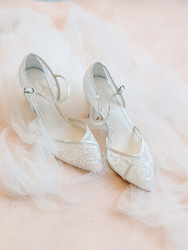 Elegant white bridal shoes with delicate lace embroidery and ankle straps, placed on soft, flowing tulle fabric.