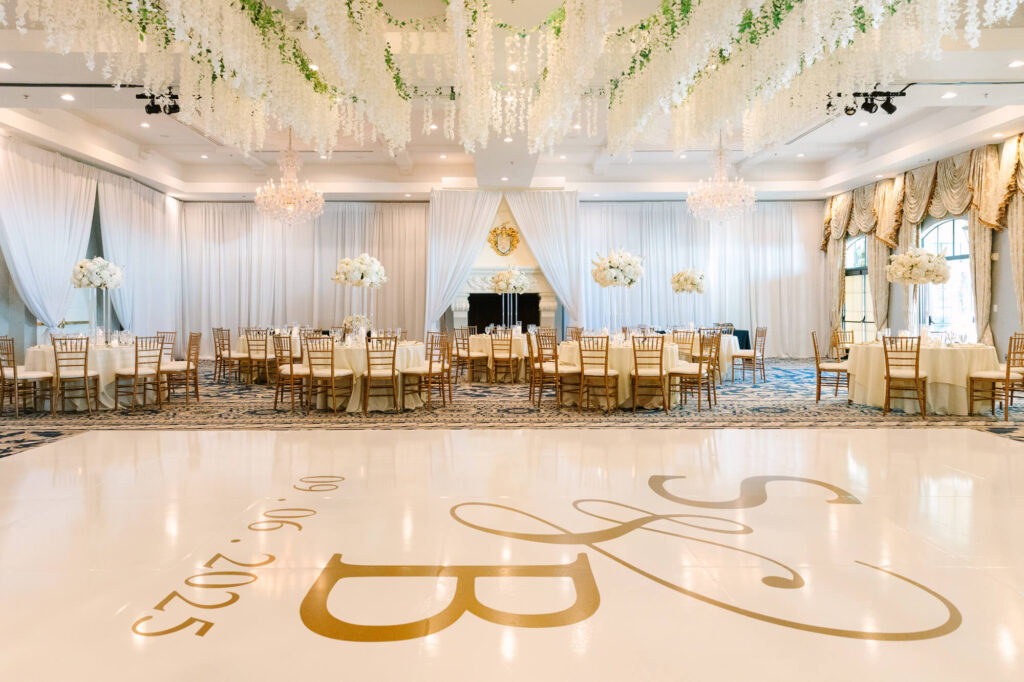 Elegant wedding reception hall with chandeliers, white floral ceiling decor, round tables with white floral centerpieces, and initials “S&B” on the floor.