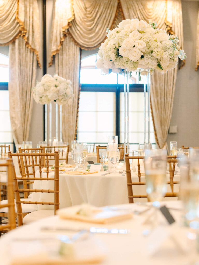Elegant wedding reception with round tables, tall floral centerpieces featuring white roses, gold chairs, soft lighting, and draped curtains in the background.